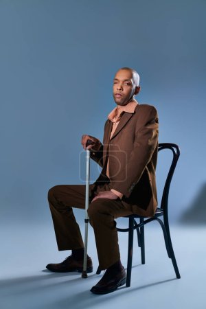diversidad e inclusión, discapacidad física, hombre africano con miastenia gravis sentado en la silla y mirando a la cámara sobre fondo azul, apoyado en bastón, dificultad para caminar
