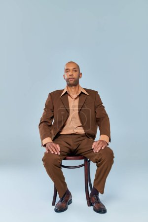 real people, full length of bold african american man with myasthenia gravis sitting on wooden chair on grey background, dark skinned person in suit looking at camera, diversity and inclusion 