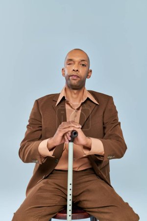Behinderter, mutiger afrikanisch-amerikanischer Mann mit Myasthenia gravis auf Holzstuhl auf grauem Hintergrund sitzend, dunkelhäutige Person im Anzug, angelehnt an Gehstock, Vielfalt und Inklusion 