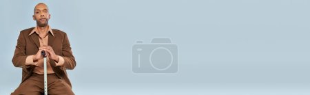 Behinderter, mutiger afrikanisch-amerikanischer Mann mit Myasthenia gravis auf Holzstuhl vor grauem Hintergrund sitzend, dunkelhäutige Person im Anzug, angelehnt an Gehstock, Vielfalt und Inklusion, Transparent 