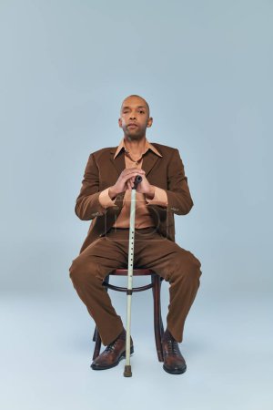 Invalidität, volle Körperlänge eines kräftigen afrikanisch-amerikanischen Mannes mit Myasthenia-Gravis-Syndrom, der auf einem Stuhl auf grauem Hintergrund sitzt, dunkelhäutige Person im Anzug, angelehnt an einen Gehstock, Vielfalt und Inklusion 