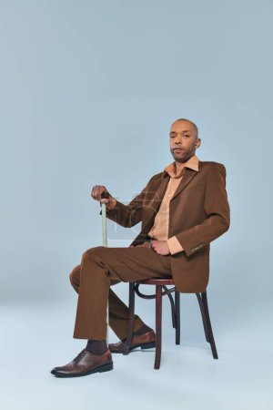 ptosis syndrome, full length of bold african american man with myasthenia gravis sitting on chair on grey background, dark skinned person in suit leaning on walking cane, diversity and inclusion 