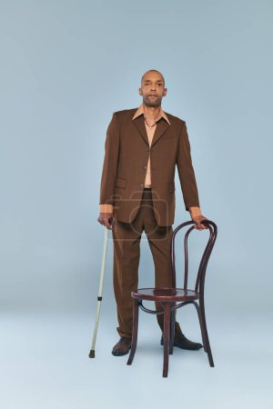 Téléchargez les photos : Syndrome de ptose, pleine longueur de gras homme afro-américain avec myasthénie gravis debout près de la chaise sur fond gris, personne à la peau foncée en costume avec canne à pied, la diversité et l'inclusion - en image libre de droit