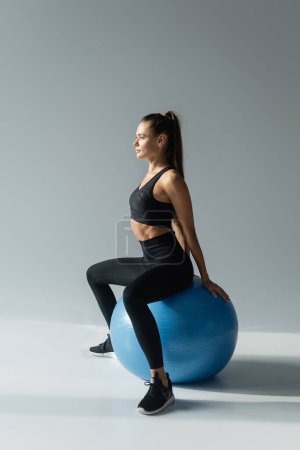 deportista en forma de sujetador deportivo negro y leggings sentado en la pelota de fitness sobre fondo gris 