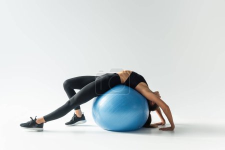 Brunette woman in sportswear stretching on fitness ball and training on white, athletic concept