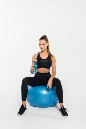 sportswoman in active wear holding sports bottle and sitting on fitness ball on white background 