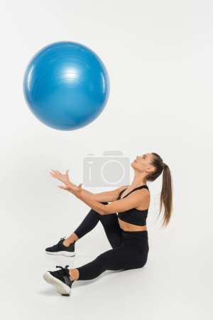 mujer en traje deportivo negro lanzando pelota de fitness y sentado sobre fondo blanco, concepto atlético
