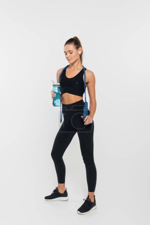 sportswoman in active wear using earphone, holding sports bottle and jump rope on white background 