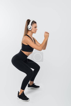 sportswoman in headphones and sportswear doing squats on white background, wellness concept