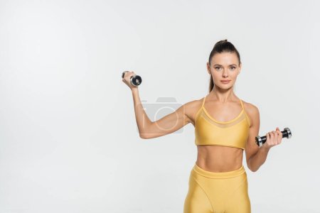 Sportlerin in Fitnessbekleidung blickt in die Kamera, Training mit Hanteln isoliert auf weiß 