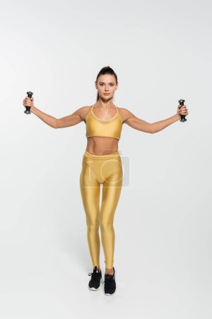mujer en entrenamiento de ropa de fitness con mancuernas y mirando a la cámara sobre fondo blanco 