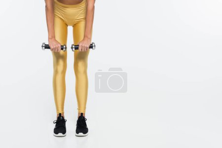 Vista recortada de la deportista en forma en entrenamiento de polainas amarillas con pesas sobre fondo blanco 