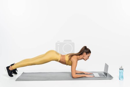 Foto de E-sports, mujer haciendo tablón en la estera de la aptitud, fondo blanco, concepto de la actividad física, ordenador portátil - Imagen libre de derechos