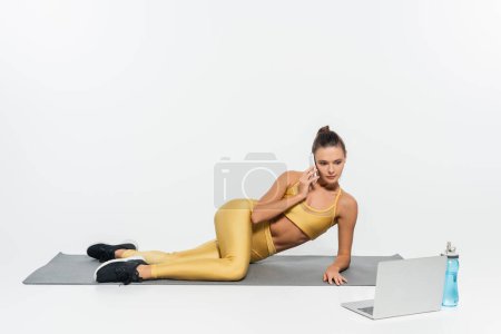 woman talking on smartphone, sitting near laptop and sports bottle on fitness mat, white background 