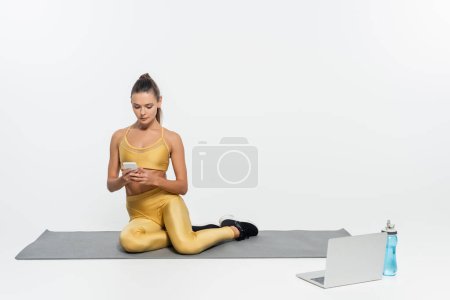 Téléchargez les photos : Femme utilisant smartphone et assis o tapis de fitness près d'un ordinateur portable sur fond blanc, l'activité physique - en image libre de droit