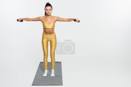 brunette sportswoman in sportswear training with dumbbells on fitness mat on white background