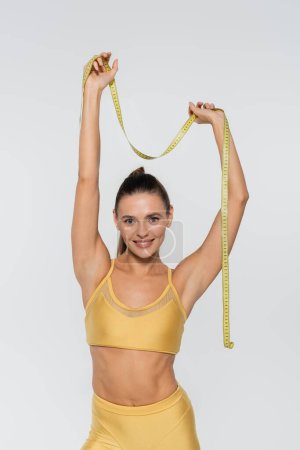 weight loss, happy sportswoman with toned body holding measuring tape above head on white background