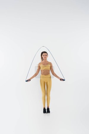 Foto de Longitud completa de la deportista morena en ropa deportiva amarilla sosteniendo saltar la cuerda y el entrenamiento en blanco - Imagen libre de derechos
