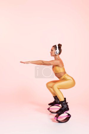 Gleichgewicht, Frau in Kangoo-Springschuhen und drahtlosen Kopfhörern, die auf rosa Hintergrund trainieren, Kniebeugen