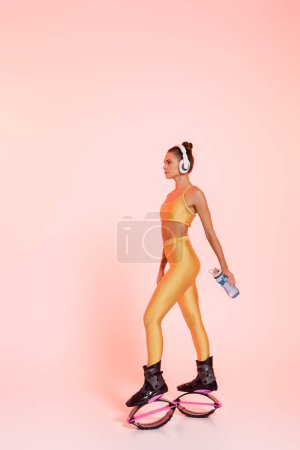 Photo for Woman in kangoo jumping shoes and wireless headphones holding sports bottle with water on pink - Royalty Free Image