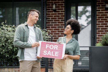 glückliche multiethnische Immobilienmakler, die zum Verkauf stehende Schilder halten und einander in der Nähe des Hauses anschauen