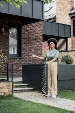 Téléchargez les photos : Heureux courtier immobilier afro-américain pointant avec la main et montrant chalet de la ville - en image libre de droit