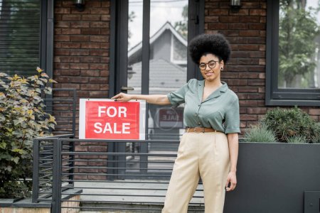 Téléchargez les photos : Agent immobilier afro-américain optimiste debout avec à vendre enseigne près de la clôture du chalet - en image libre de droit