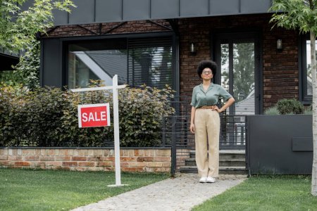 agente inmobiliario afroamericano feliz con la mano en la cadera cerca del edificio para la venta en la ciudad