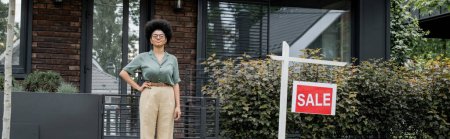 agente inmobiliario afroamericano complacido con la mano en la cadera cerca de casa urbana para la venta, pancarta