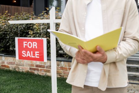 Téléchargez les photos : Vue recadrée de l'agent immobilier debout avec dossier près de panneau à vendre en face de la maison de la ville - en image libre de droit