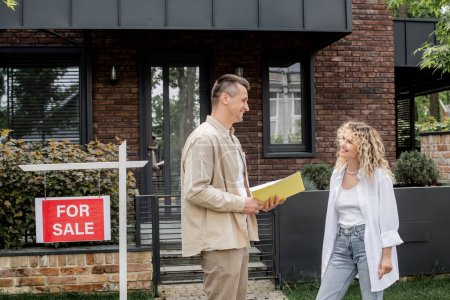 fröhlicher Immobilienmakler mit Folder im Gespräch mit Frau in der Nähe moderner Hütte in der Stadt