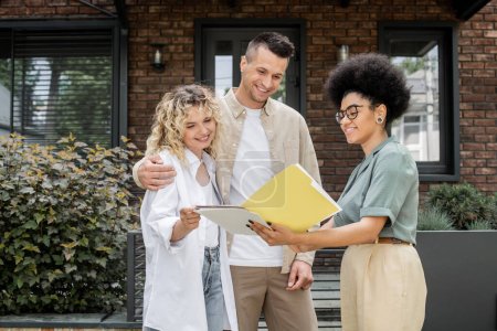 african american property realtor showing documents to happy couple near new modern cottage