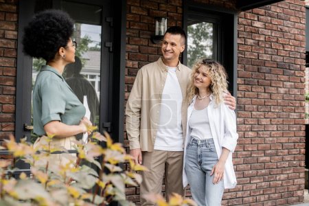 pleased couple embracing while talking with african american realtor near new cottage on street