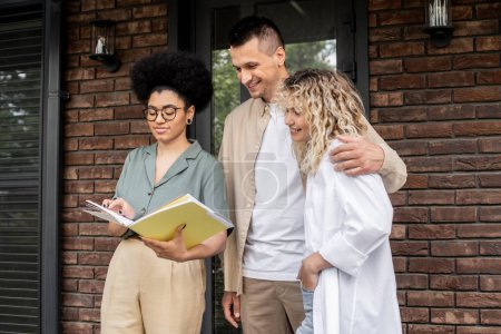 african american real estate agent showing documents to delighters buyers hugging near new house