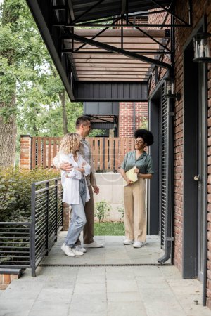 sonriente agente de bienes raíces afroamericano con carpeta hablando con un par de nuevos propietarios cerca de la casa