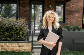 cheerful blonde real estate agent holding documents and looking at camera near house on city street Stickers #664924340