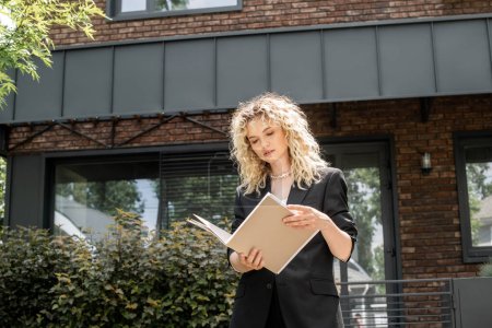 stylish blonde real estate agent looking at documents in folder near contemporary urban building