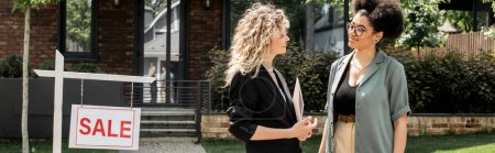 Téléchargez les photos : Souriant afro-américaine parler à blonde agent immobilier près de chalet à vendre, bannière - en image libre de droit