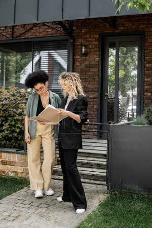 Téléchargez les photos : Agent immobilier blonde montrant des documents à un client afro-américain près d'une maison moderne à vendre - en image libre de droit