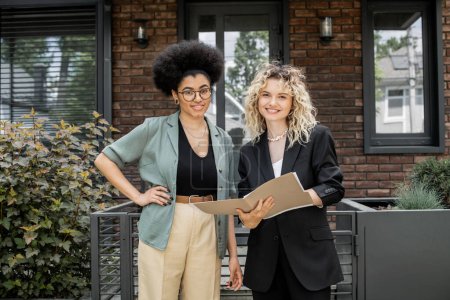 Téléchargez les photos : Heureux agent immobilier avec dossier et afro-américaine femme avec la main sur la hanche regardant caméra près de la maison - en image libre de droit