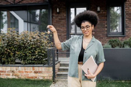 erfolgreiche afrikanisch-amerikanische Immobilienmaklerin mit Schlüssel und Ordner lächelt in die Kamera in der Nähe von neuem Ferienhaus