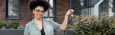 smiling african american real estate agent in eyeglasses showing key from new house outdoors, banner