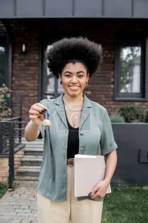 Téléchargez les photos : Heureux et réussi courtier immobilier afro-américain avec dossier détenant clé près de nouveau chalet - en image libre de droit