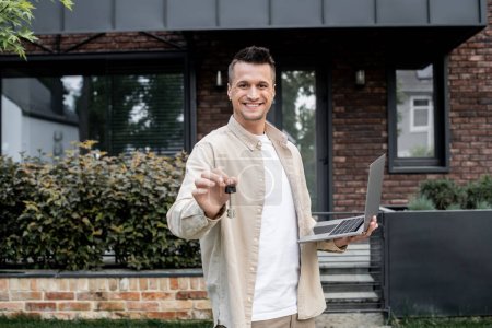 fröhlicher Immobilienmakler mit Laptop zeigt Schlüssel aus neuem Haus, während er draußen steht