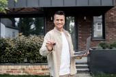 cheerful real estate agent with laptop showing key from new house while standing outdoors t-shirt #664924678
