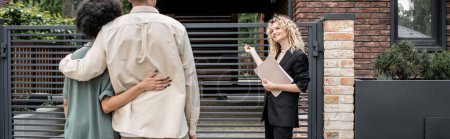 Téléchargez les photos : Agent immobilier blonde positive avec dossier pointant vers une nouvelle maison près d'un couple multiculturel, bannière - en image libre de droit