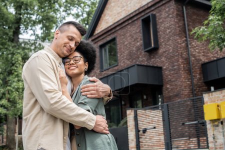 alegre pareja multiétnica abrazando delante de la nueva casa de campo propia en la calle urbana, pancarta