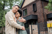 joyful multiethnic couple embracing in front of new own cottage on urban street, banner mug #664924804