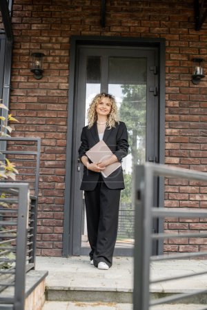 full length of stylish and confident realtor standing with folder near new house in city