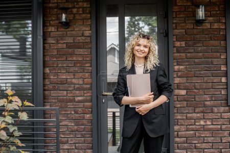 blonde and attractive real estate agent holding folder and smiling at camera near city cottage